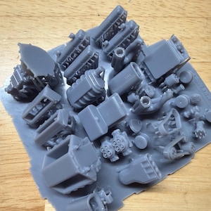 May include: A collection of gray miniature engine parts, including blocks, cylinders, and other components, are arranged on a light gray sheet. The parts are likely for model building or a similar hobby. The background is a light brown wooden surface.