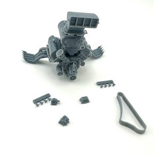 May include: A gray model of a car engine with multiple components, including an air intake, exhaust manifolds, and smaller detached parts. The engine is designed with intricate details, showcasing its mechanical complexity.