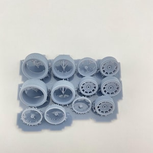 Puede incluir: Una colección de pequeñas piezas de modelo de color gris claro, que incluyen componentes de motor cilíndricos y estructuras en forma de rueda. Las piezas están dispuestas sobre una base rectangular, probablemente para un kit de modelo.