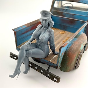 Peut inclure: Figurine grise d'une femme en uniforme et casquette de style militaire, assise sur le hayon d'un pick-up bleu et marron vieilli. La figurine est détaillée avec des talons hauts et une tenue moulante.
