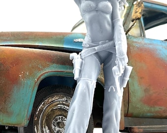 May include: A gray figurine of a woman in a cowboy hat and fringed sleeves, leaning against a weathered, rusty blue truck. She wears a bikini top, pants, and a gun belt with holsters. The figure is detailed and stands out against the white background.