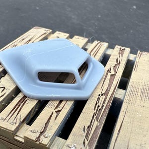 Könnte beinhalten: Ein grauer Kunststoff-Motorhauben-Scoop mit zwei rechteckigen Lüftungsschlitzen. Der Scoop liegt auf einer Holzpalette.