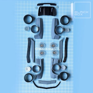 May include: An overhead shot of disassembled gray and black car parts arranged on a blue grid surface. The parts include wheels, body panels, and other components, suggesting a model car kit or repair project. The text "BLACK BOX" is visible.