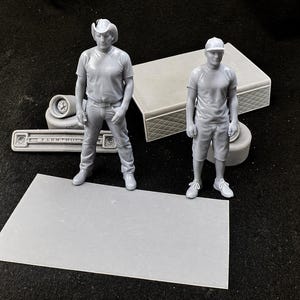 May include: Two gray miniature figures of people, one wearing a cowboy hat and the other a baseball cap. They stand near model truck parts, including a front grill with the text "FARM TRUCK". A rectangular base and other model parts are also present.