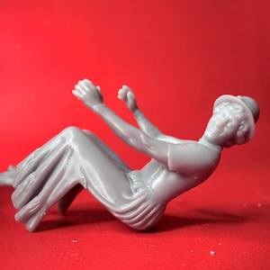 May include: A gray plastic figurine of a person in a seated position, wearing a hat and long skirt. The figure's arms are raised, and the details of the clothing are visible. The background is a solid red.