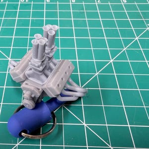 May include: A small grey engine model on a blue keyring. The engine features multiple exhaust pipes and is set against a green grid background. The keyring is made of metal.