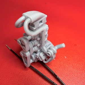 May include: A small, gray model of an engine, featuring intricate details of its components. The model includes curved pipes, a rectangular top, and various mechanical parts. The engine is positioned on a red surface, with black tweezers in the foreground.