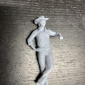 May include: A light gray figurine of a person in a cowboy hat, button-down shirt, jeans, and boots. The figure is leaning against an unseen object, with one arm bent at the elbow and the other resting on the hip. The figure has a belt with a large buckle.