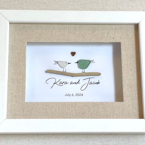 May include: A framed artwork featuring two pebble birds perched on a driftwood branch. The birds are white and green, with a small heart above them. The names "Kara and Jacob" and the date "July 6, 2024" are written below.