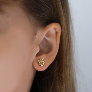 Puede incluir: Primer plano de una oreja con un pendiente de botón dorado con forma de huella de pata. El pendiente es pequeño y detallado, mostrando la forma de una pata. El fondo es de un color suave y neutro, resaltando el diseño.