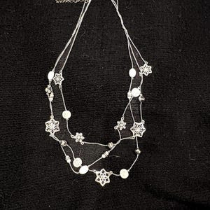 May include: A silver-toned, multi-strand necklace featuring snowflake pendants and white, round beads. The necklace has a delicate chain and is designed with three layers. The background is a dark, textured surface.