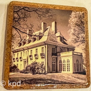 May include: Square coaster featuring a sepia-toned image of a large house with multiple windows, balconies, and chimneys. The house is surrounded by trees. The coaster has a textured, brown border. The text "© kpd" is in the lower left corner.