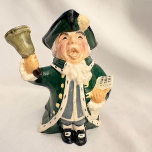 May include: A ceramic figure of a town crier in a green coat, black hat, and white ruffled collar, holding a bell and a scroll. The figure has a smiling face with rosy cheeks and is wearing black shoes. The scroll reads "OYEZ! OYEZ! OYEZ!"