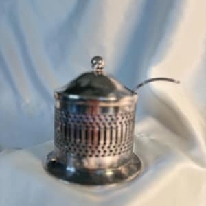 May include: A silver-colored sugar bowl with a lid and a small spoon. The bowl has a perforated design around the sides and a round base. The lid has a small ball-shaped handle.