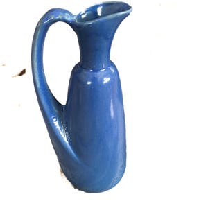 May include: A blue ceramic pitcher with a curved handle and a flared spout. The pitcher has a smooth, glossy finish and a rounded base. The handle is gracefully arched, and the overall design is elegant and functional.