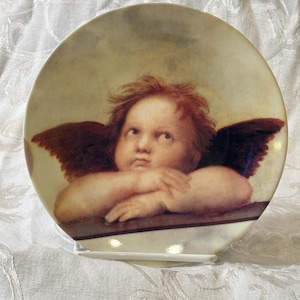 May include: A round ceramic decorative plate featuring a detailed reproduction of a cherub with reddish-brown hair and dark wings. The cherub is shown from the chest up, resting its chin on folded arms, with a soft, muted background color palette.