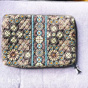 May include: A quilted, rectangular case with a floral pattern in brown, teal, yellow, and pink. The case has a zipper closure and a decorative pull. The design features a central vertical stripe with floral motifs.