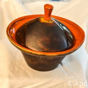 May include: A brown ceramic serving dish with a lid. The lid has a small knob on top and the dish has a wide orange rim.