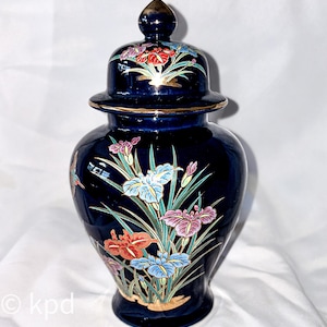 May include: A dark blue ceramic ginger jar with a lid, featuring a floral design of irises in shades of red, pink, blue, and white. The jar has gold trim and a gold finial on the lid. The overall design is a classic, decorative style.