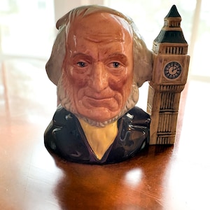 May include: A ceramic figurine of a distinguished-looking man with white hair and a black coat, featuring a Big Ben clock tower on the side. The figurine has a detailed face and is painted in muted tones.