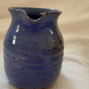 May include: A handmade, blue ceramic creamer with a spout. The creamer has a rounded body and a textured surface with dark speckles. The rim is slightly darker in color. The creamer is approximately 4 inches tall.