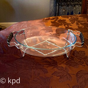May include: An oval glass baking dish with a silver metal stand and black handles. The dish is clear, allowing visibility of its contents. The stand provides support and elevation. The dish is designed for oven use.