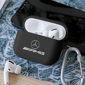 Puede incluir: Funda protectora negra para auriculares inalámbricos con el logotipo AMG en blanco. La funda está abierta, revelando auriculares blancos. Un mosquetón plateado está adjunto. Un smartphone está en el fondo.