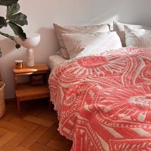 Könnte beinhalten: Ein Bett ist mit einer korallenfarbenen und weißen gemusterten Tagesdecke bedeckt, mit weißen Kissen. Ein Nachttisch aus Holz mit einer Lampe und einem kleinen goldenen Behälter steht neben dem Bett. Der Raum hat Parkettböden und eine große Pflanze.