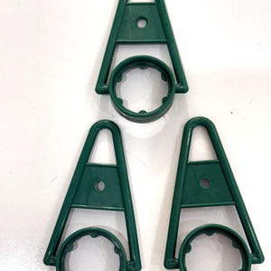 May include: Three dark green plastic triangular hangers, each featuring a central circular opening with small interior notches and a smaller circular hole in the upper section. These items have a rigid frame and a smooth, molded finish.