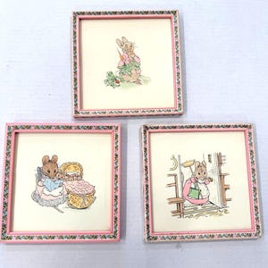 May include: Three framed illustrations featuring rabbit characters in various scenes. Each square frame has a pink border with a floral pattern. The illustrations depict rabbits with carrots, in a crib, and sweeping.