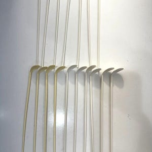 May include: A collection of fourteen off-white plastic stirrers, each featuring a long, slender handle and a small, rounded, angled paddle at the top. These uniform utensils are arranged in two horizontal rows, showcasing a simple, functional design.