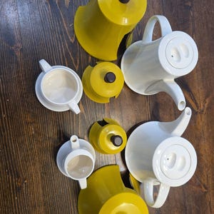 Puede incluir: Un juego de té compuesto por dos teteras de cerámica blanca con tapas a juego, dos tazas de cerámica blanca y tres cubiertas de color amarillo mostaza con pomos de color marrón oscuro, dispuesto en una vista cenital.