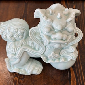 May include: A light blue ceramic guardian lion statue. The mythical creature has a detailed design with a fierce expression, curly mane, and a ball under its paw. The statue is on a dark wooden surface.