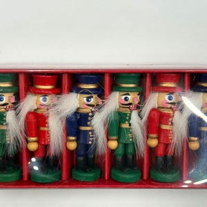 Könnte beinhalten: Ein Set aus sechs hölzernen Nussknacker-Figuren in einer roten rechteckigen Schachtel. Die Nussknacker tragen grüne, rote und blaue Uniformen mit goldenen Akzenten, schwarzen Stiefeln und weißem Kunsthaar. Jede Figur steht auf einem runden Sockel.