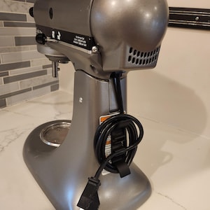 Pode incluir: Um misturador de pedestal prateado com um cabo preto enrolado em torno da base. O misturador tem uma tigela prateada e uma cabeça prateada com um botão preto. O misturador está em uma bancada branca.