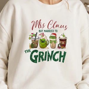 K&ouml;nnte beinhalten: Wei&szlig;er Pullover mit einer gr&uuml;nen und roten Grafik, auf der "Mrs. Claus, aber verheiratet mit dem Grinch" steht. Die Grafik zeigt vier Cartoon-Becher mit gr&uuml;nen und roten Designs.