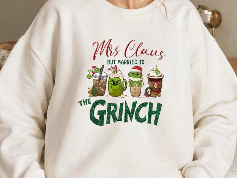 K&ouml;nnte beinhalten: Wei&szlig;er Pullover mit einer gr&uuml;nen und roten Grafik, auf der "Mrs. Claus, aber verheiratet mit dem Grinch" steht. Die Grafik zeigt vier Cartoon-Becher mit gr&uuml;nen und roten Designs.