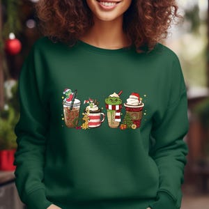 K&ouml;nnte beinhalten: Gr&uuml;nes Sweatshirt mit einem festlichen Design aus vier verschiedenen weihnachtlichen Kaffeegetr&auml;nken. Jedes Getr&auml;nk hat einen einzigartigen Charakter obenauf, darunter ein Schneemann, ein Rentier, ein Grinch und ein Weihnachtsmann.