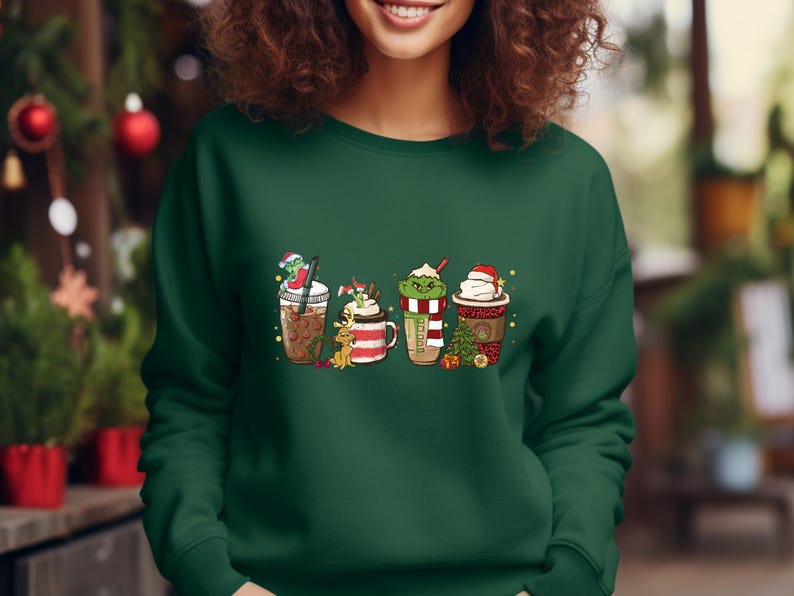 K&ouml;nnte beinhalten: Gr&uuml;nes Sweatshirt mit einem festlichen Design aus vier verschiedenen weihnachtlichen Kaffeegetr&auml;nken. Jedes Getr&auml;nk hat einen einzigartigen Charakter obenauf, darunter ein Schneemann, ein Rentier, ein Grinch und ein Weihnachtsmann.