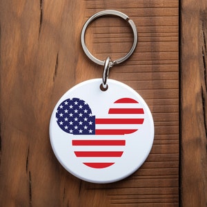 May include: A white round keychain with a red, white, and blue American flag design in the shape of Mickey Mouse's head.