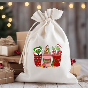 K&ouml;nnte beinhalten: Eine wei&szlig;e Stofftasche mit Kordelzug und einem weihnachtlichen Design mit drei Figuren in roten und gr&uuml;nen Bechern. Die Figuren sind der Grinch, Cindy Lou Who und Max der Hund. Das Design enth&auml;lt rote und gr&uuml;ne Ornamente, Zuckerstangen und einen gr&uuml;nen Baum.