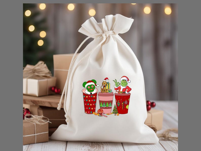 K&ouml;nnte beinhalten: Eine wei&szlig;e Stofftasche mit Kordelzug und einem weihnachtlichen Design mit drei Figuren in roten und gr&uuml;nen Bechern. Die Figuren sind der Grinch, Cindy Lou Who und Max der Hund. Das Design enth&auml;lt rote und gr&uuml;ne Ornamente, Zuckerstangen und einen gr&uuml;nen Baum.