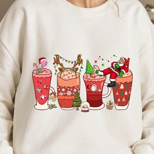 K&ouml;nnte beinhalten: Wei&szlig;er Pullover mit einem weihnachtlichen Design mit vier Bechern, gef&uuml;llt mit hei&szlig;er Schokolade oder Kaffee. Jeder Becher hat einen anderen festlichen Charakter oder eine andere Dekoration, darunter den Weihnachtsmann, ein Rentier, einen Weihnachtsbaum und einen Zuckerstock.