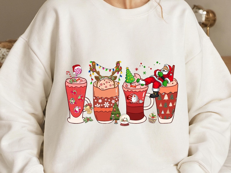 K&ouml;nnte beinhalten: Wei&szlig;er Pullover mit einem weihnachtlichen Design mit vier Bechern, gef&uuml;llt mit hei&szlig;er Schokolade oder Kaffee. Jeder Becher hat einen anderen festlichen Charakter oder eine andere Dekoration, darunter den Weihnachtsmann, ein Rentier, einen Weihnachtsbaum und einen Zuckerstock.