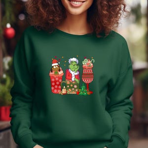 K&ouml;nnte beinhalten: Gr&uuml;ner, roter und wei&szlig;er Weihnachtspullover mit einer Cartoon-Illustration des Grinch, des Weihnachtsmanns und Cindy Lou Who in Bechern. Der Pullover hat einen gr&uuml;nen Hintergrund.