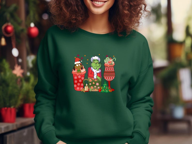 K&ouml;nnte beinhalten: Gr&uuml;ner, roter und wei&szlig;er Weihnachtspullover mit einer Cartoon-Illustration des Grinch, des Weihnachtsmanns und Cindy Lou Who in Bechern. Der Pullover hat einen gr&uuml;nen Hintergrund.