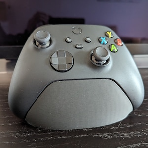 May include: A black Xbox controller with a textured surface and a black base. The controller has a directional pad, four face buttons, two analog sticks, and two triggers.