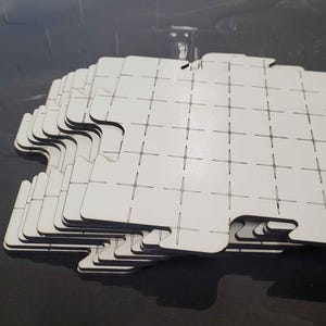 Pode incluir: Uma pilha de pisos quadrados brancos de encaixe, apresentando um padrão de grade com linhas pretas finas e pequenas marcas de cruz. Cada peça possui bordas entalhadas projetadas para conectar-se com as peças adjacentes.