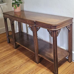 May include: A long, rectangular wooden console table with a rich brown finish. The top surface features a chevron wood grain pattern. The table has four legs with decorative corner brackets and includes a lower shelf for additional storage space.
