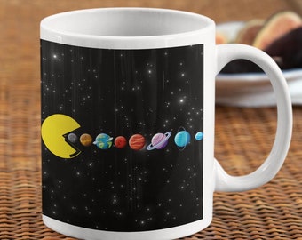 May include: A white ceramic mug with a black background and a yellow Pac-Man character eating the planets of the solar system. The planets are in order from the sun: Mercury, Venus, Earth, Mars, Jupiter, Saturn, Uranus, and Neptune.